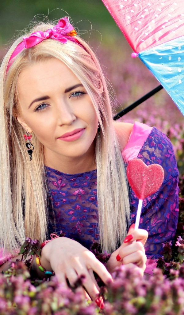 Молодая девушка блондинка лежит на поляне с сиреневыми цветами под разноцветным зонтом