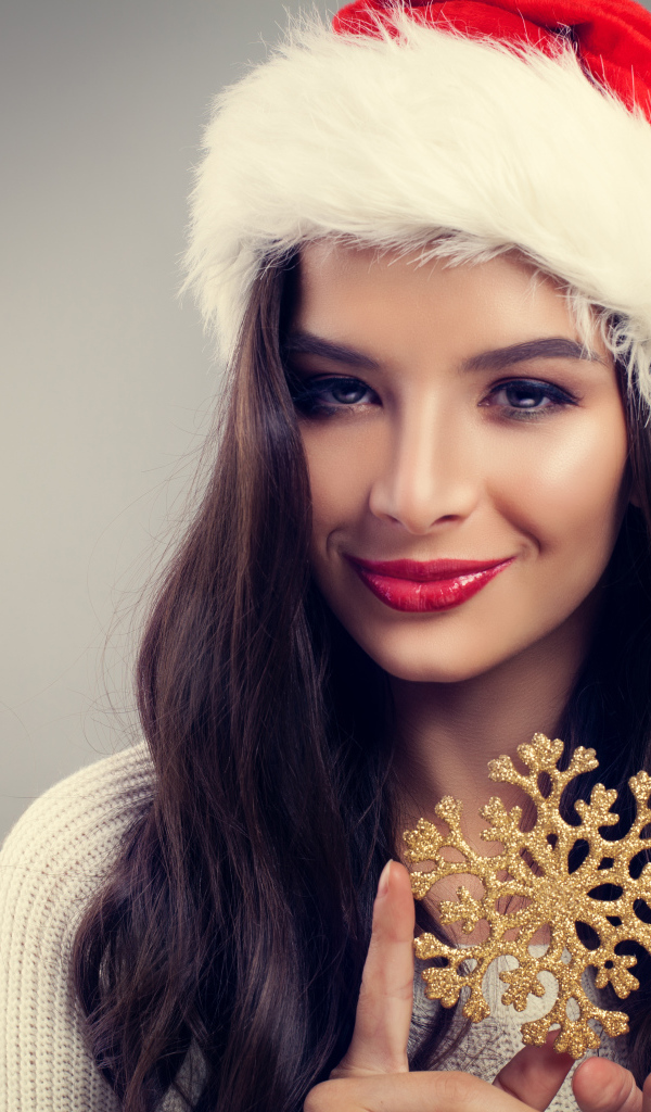 Красивая яркая брюнетка в новогодней шапке со снежинкой в руках на сером фоне