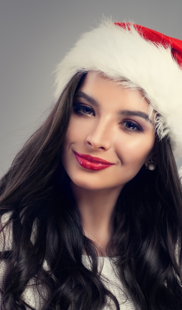 Красивая длинноволосая девушка брюнетка в шапке деда Мороза на сером фоне
