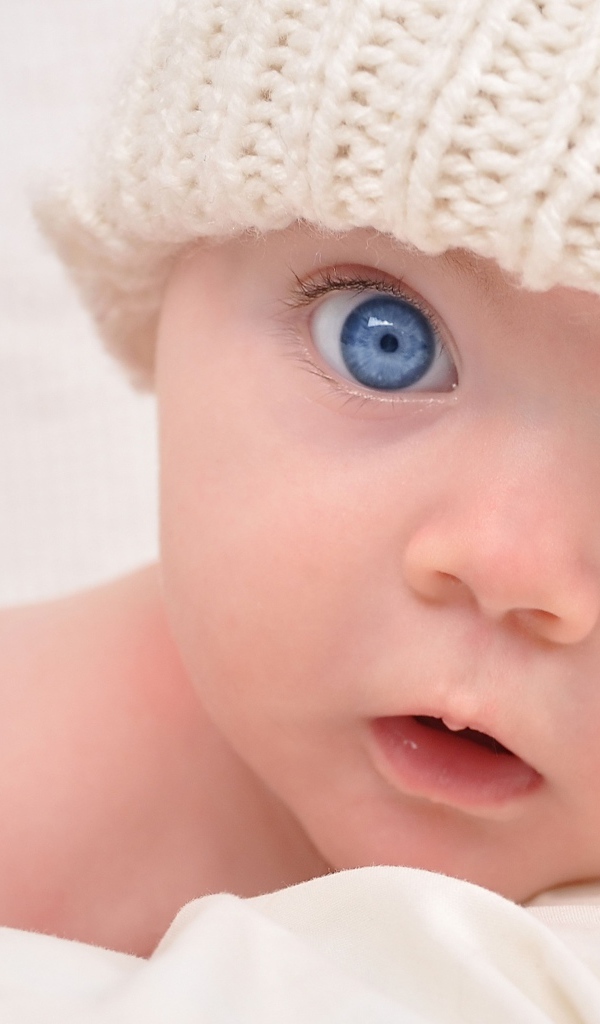 Голубоглазый малыш в белой вязаной шапке на сером фоне