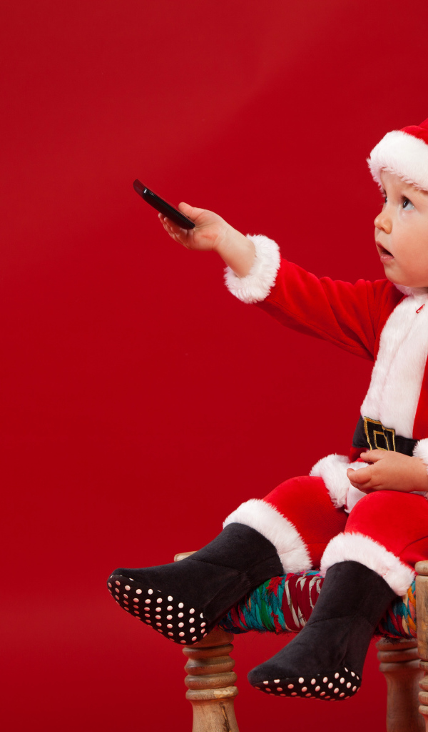 Маленький мальчик в костюме Санта Клауса сидит на стуле на красном фоне