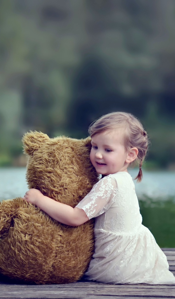 Маленькая девочка в белом платье сидит на мосту с плюшевым медведем