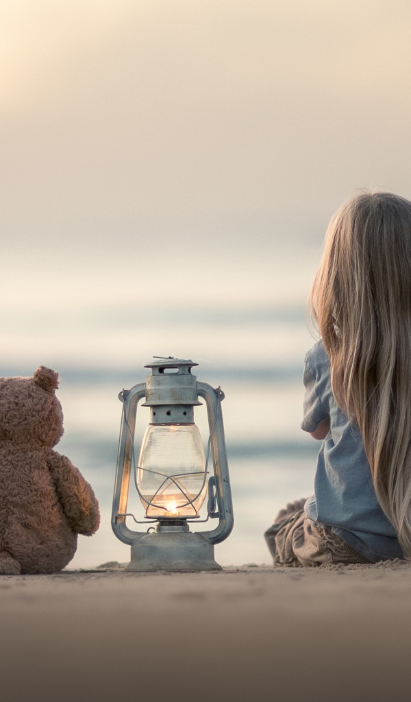 Маленькая девочка сидит на песке с плюшевым мишкой 