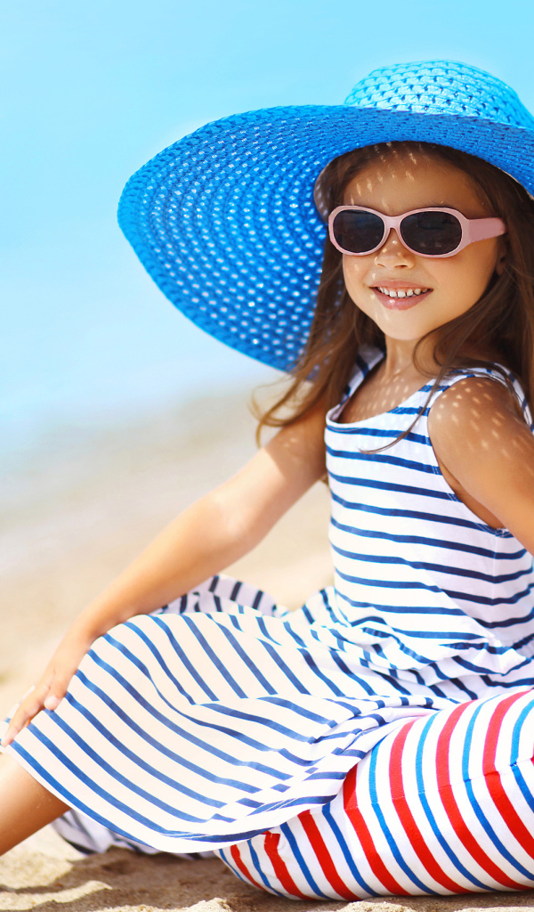 Маленькая девочка сидит на подушке на пляже в большой синей шляпе