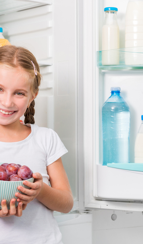 Маленькая девочка с миской со сливами в руках у холодильника 