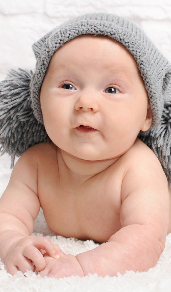 Улыбающийся грудной ребенок в серой шапке с бубонами