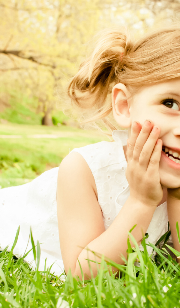 Улыбающаяся девочка в белом платье лежит на зеленой траве
