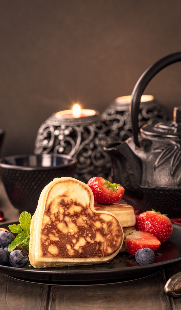 Оладьи в форме сердца с ягодами черники и клубники на столе с чаем и свечами