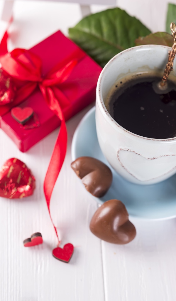 Чашка кофе на столе с шоколадными конфетами, подарком и красной розой