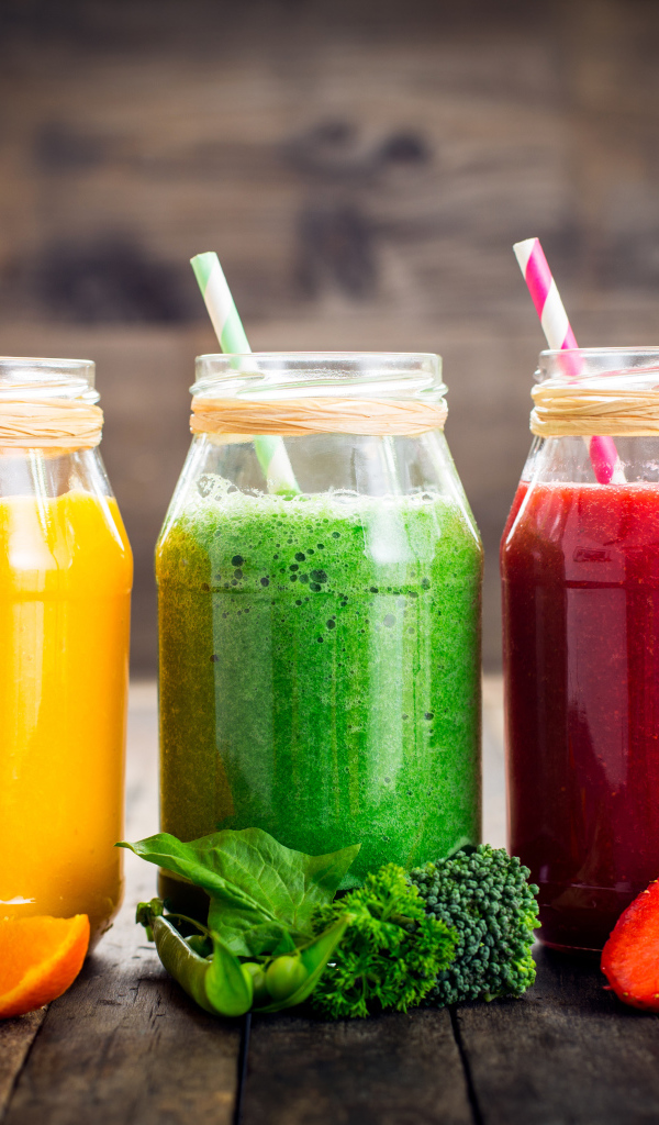 Три банки смузи с трубочкой на столе с апельсином, брокколи и клубникой