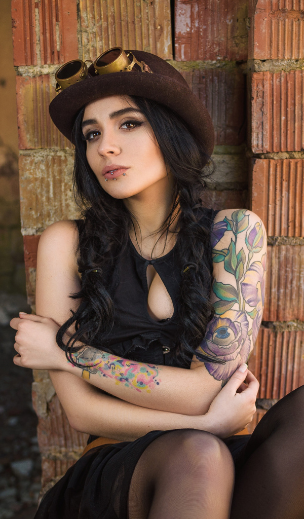 Девушка в шляпе с татуировками на руке сидит у стены