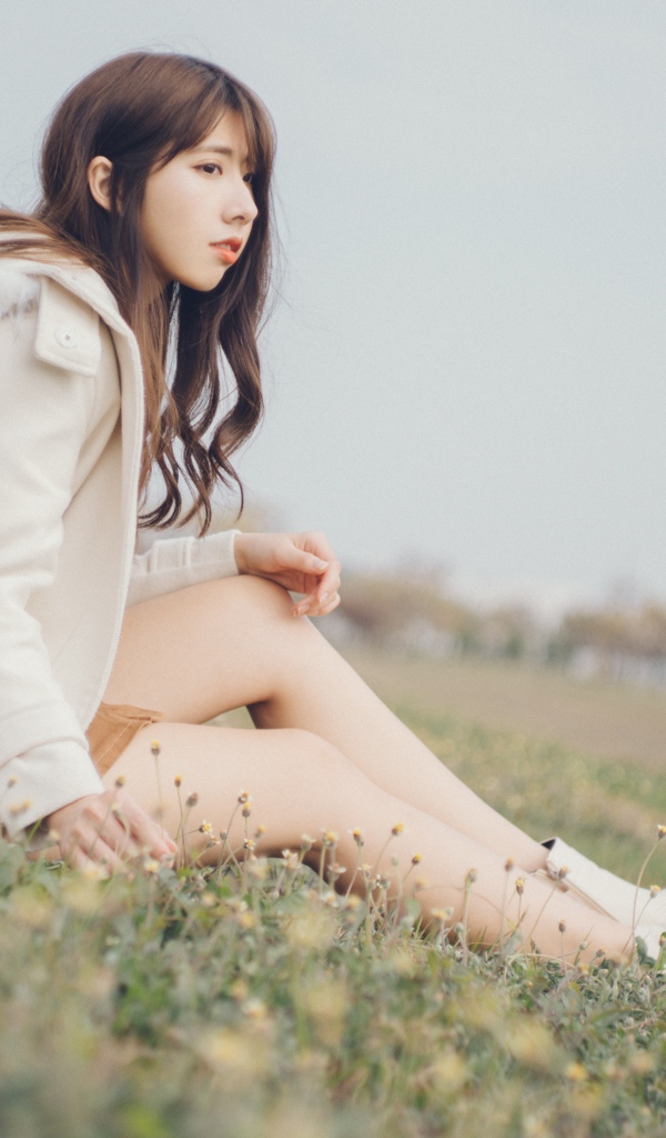 Девушка азиатка в пальто сидит на траве