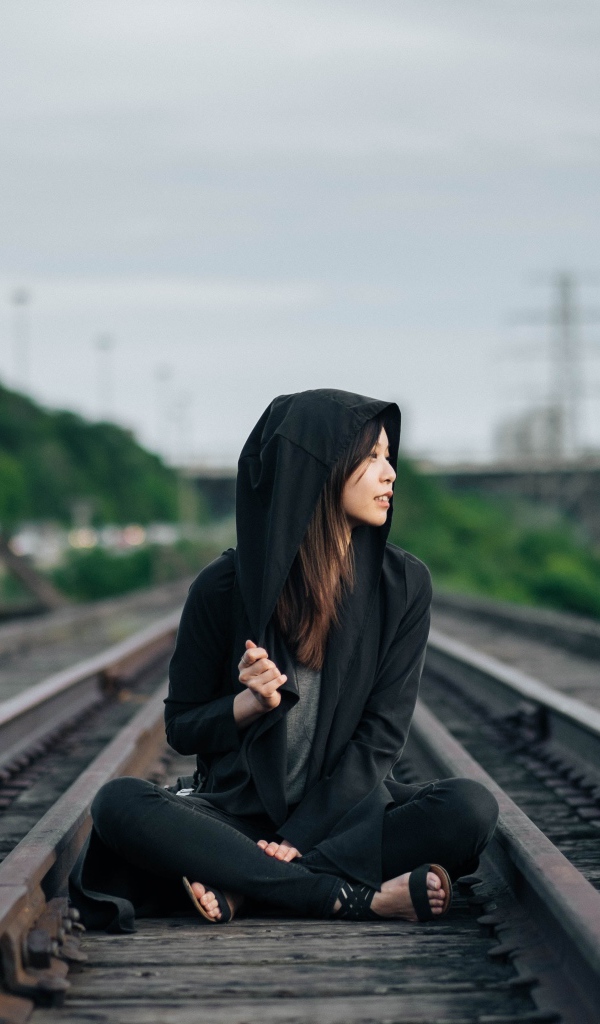Девушка азиатка в черном балахоне сидит на рельсах 