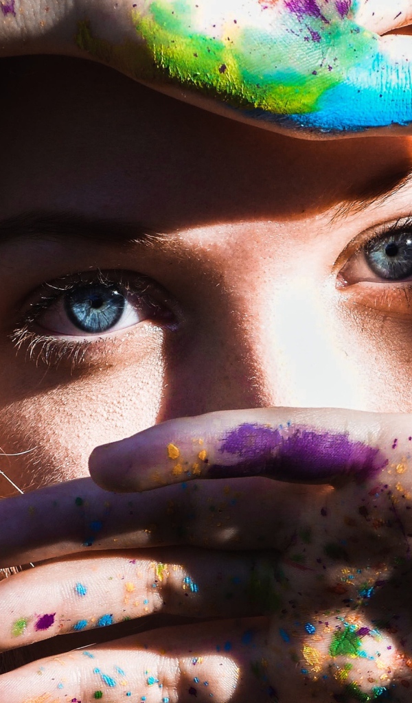 Красивая голубоглазая девушка с руками в разноцветной краске у лица