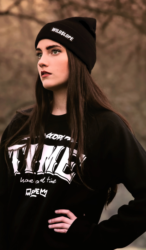 Красивая девушка в черной шапке у деревьев 