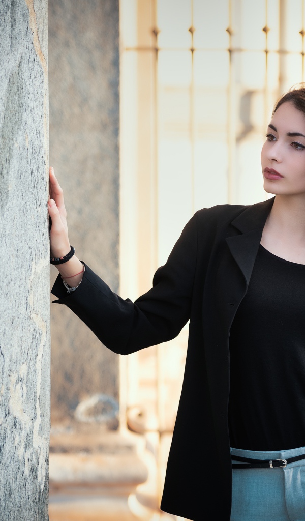 Красивая девушка в черном пиджаке стоит у стены