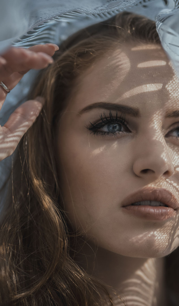 Голубоглазая девушка смотрит в окно с тюлью