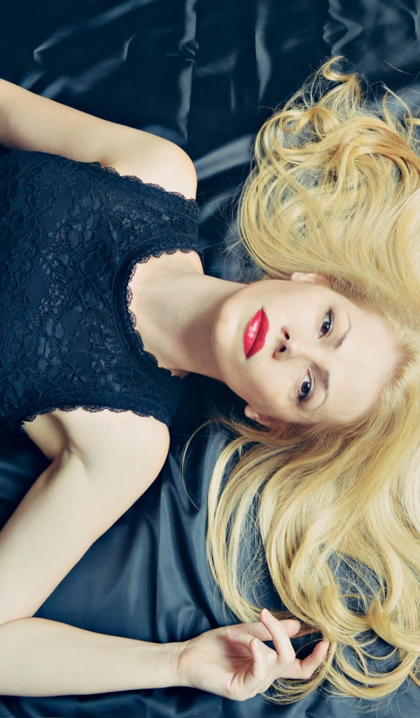 Длинноволосая блондинка в черном платье лежит на кровати