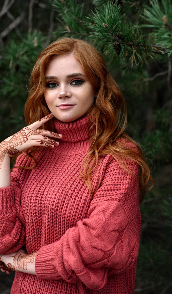 Рыжеволосая девушка в розовом свитере стоит у ели