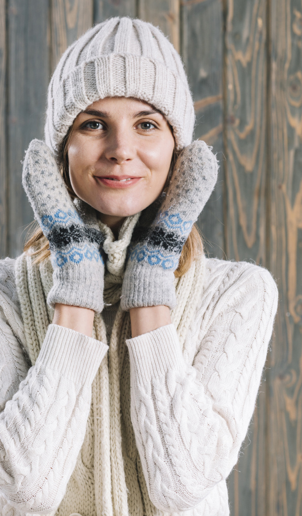 Молодая девушка на шапке, варежках  и теплой шапке на фоне забора