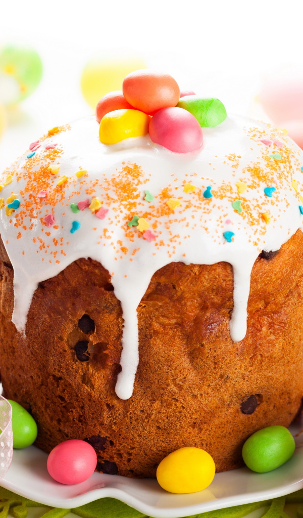 Красивый пасхальный кулич с белой глазурью и конфетами на столе с яйцами