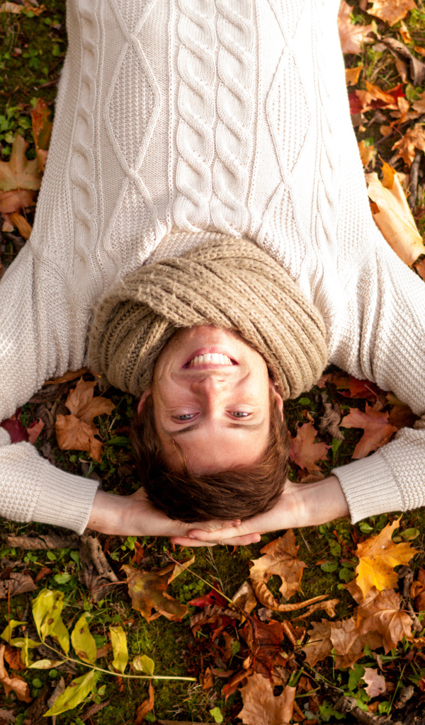 Улыбающийся мужчина в свитере лежит на опавших листьях 