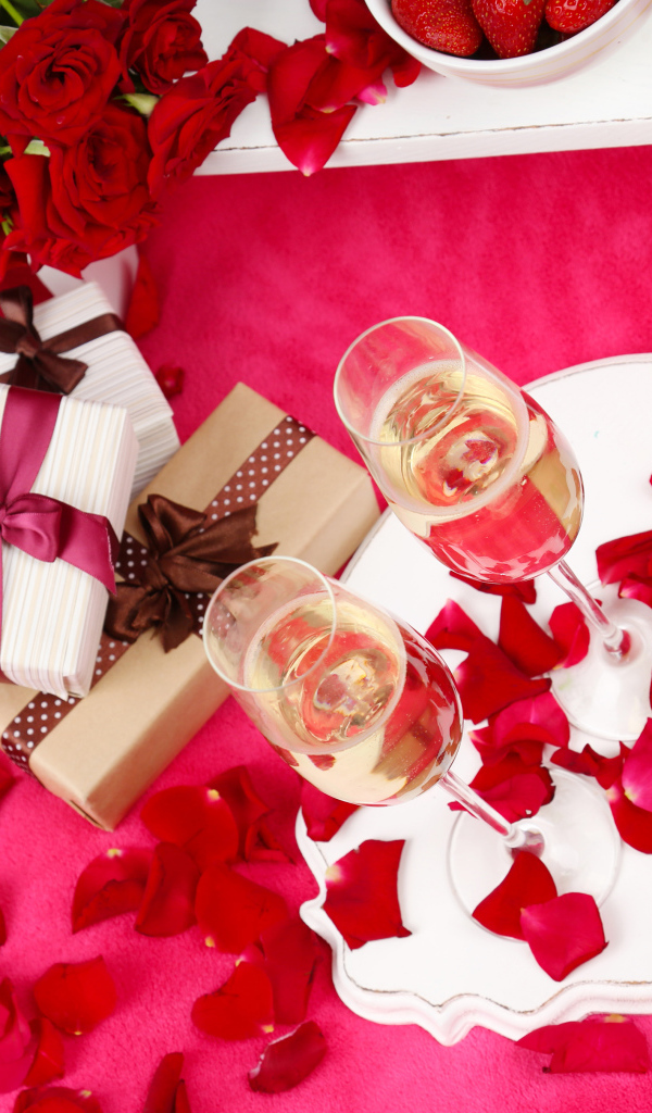 Два фужера с шампанским на красном покрывале с лепестками, подарками и букетом роз для любимой