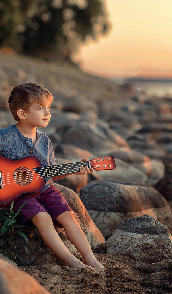 Маленький мальчик сидит с гитарой на камнях у моря 