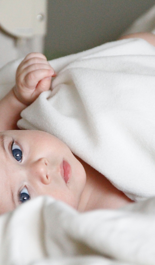 Маленький грудной голубоглазый ребенок лежит под белым покрывалом 