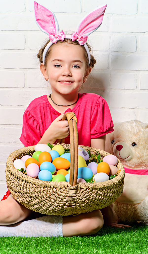 Улыбающаяся девочка сидит у стены с корзиной пасхальных яиц и игрушками