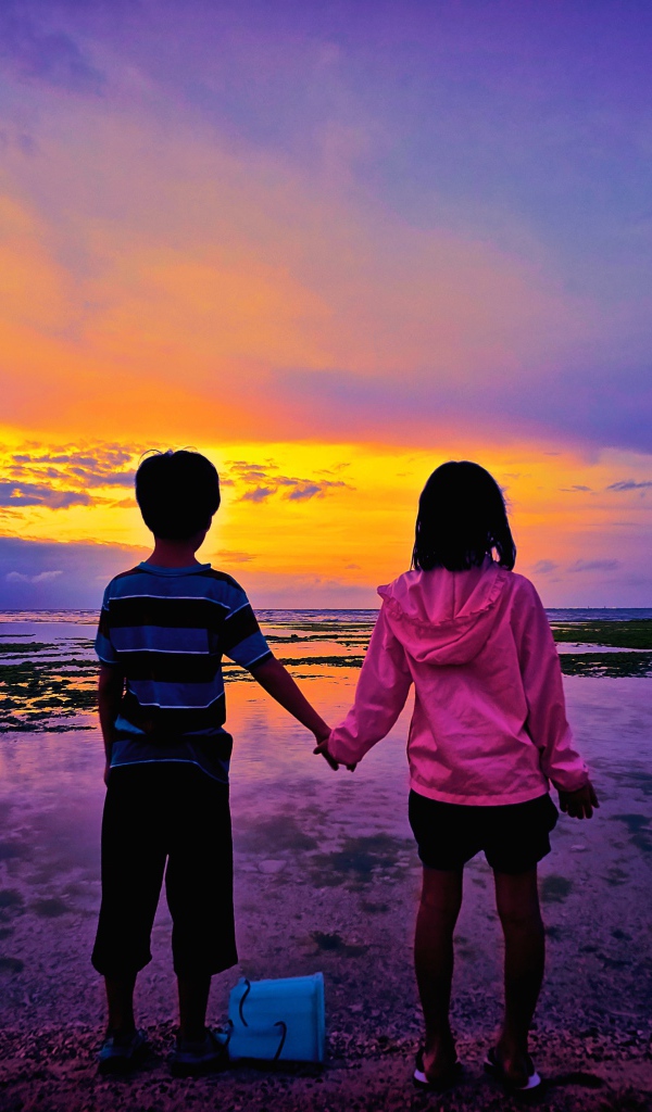 Маленькие мальчик и девочка стоят у воды на фоне заката 