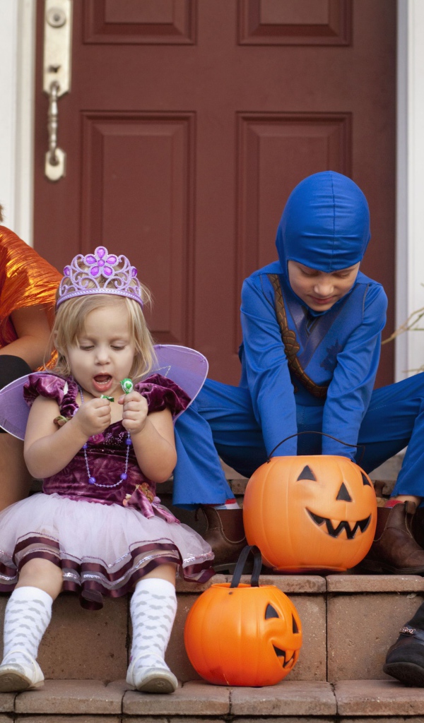 Маленькие дети в костюмах на Хэллоуин сидят на ступеньках со сладостями