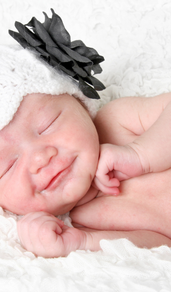 Улыбающийся спящий ребенок в белой вязаной шапке с цветком