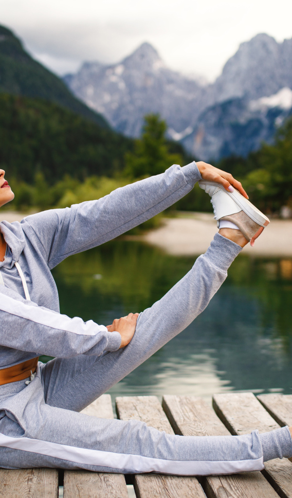 Девушка в наушниках занимается гимнастикой на мосту у озера