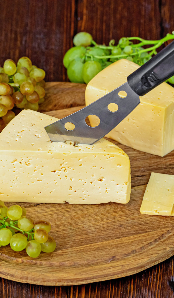 Кусок твердого сыра на столе с ножом и белым виноградом