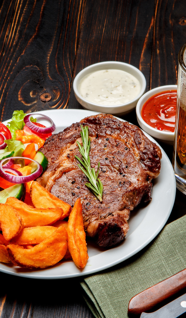 Кусок мяса на тарелке с картофелем и салатом на столе с соусом и пивом