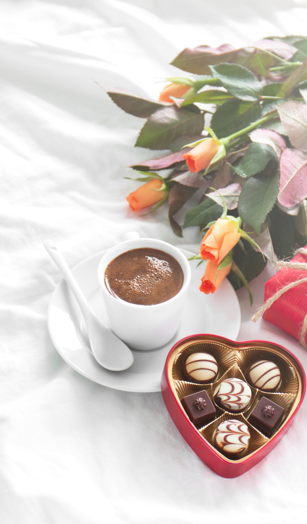 Чашка кофе на белой кровати с конфетами, подарком и букетом роз
