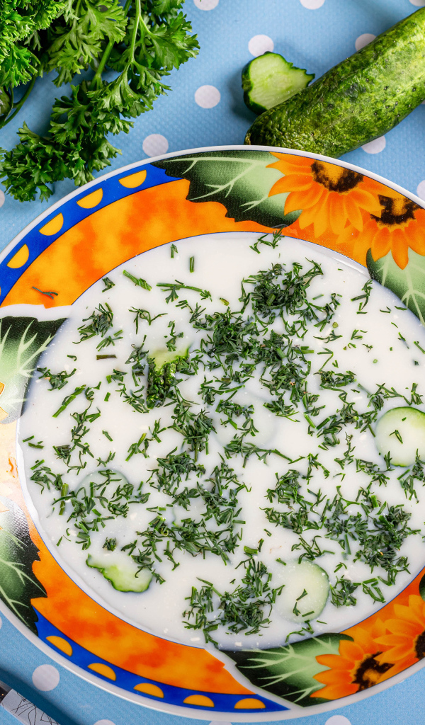 Окрошка в тарелке на столе с зеленью и огурцами