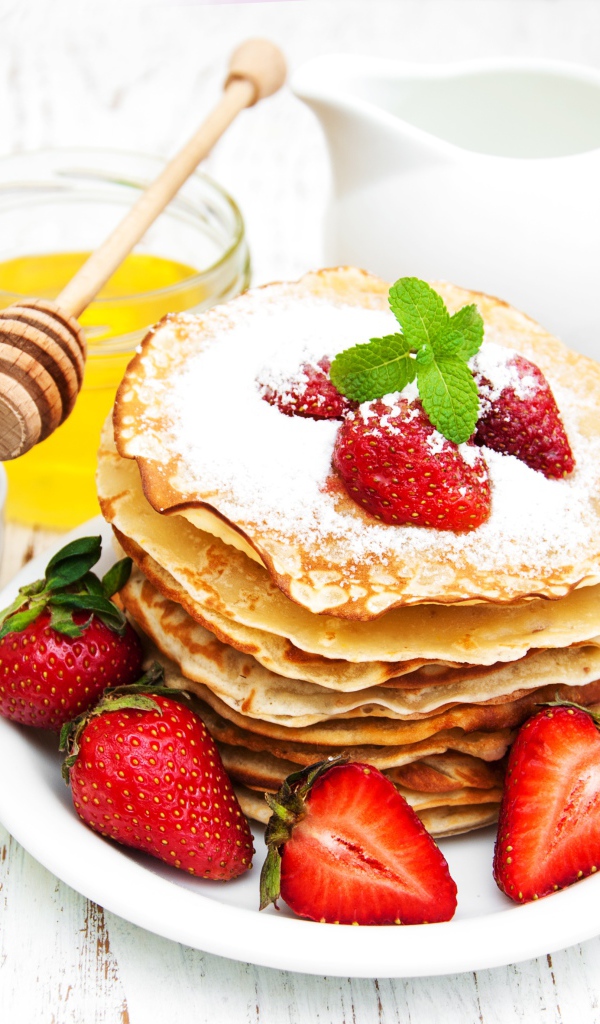 Тонкие блины с сахарной пудрой и ягодами клубники на столе с молоком и медом