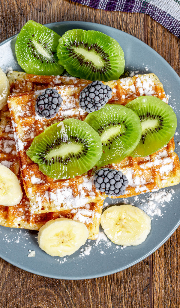 Сладкие вафли на тарелке с сахарной пудрой, бананами и киви