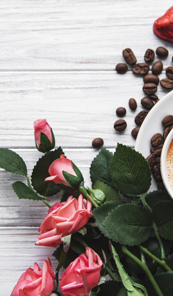 Чашка кофе с рисунком на пене на столе с букетом роз, зернами и конфетами