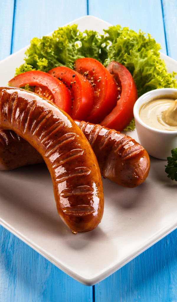 Две жареные колбаски на тарелке с помидорами, горчицей и листьями салата 