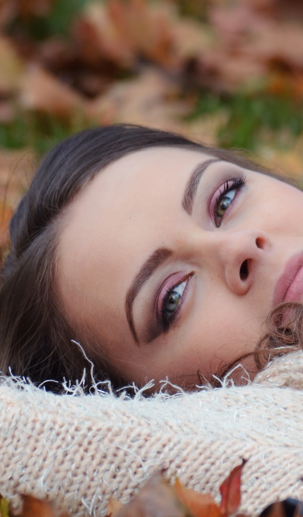 Красивая девушка в теплом свитере лежит на сухой листве 