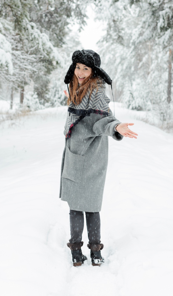 Девушка в пальто и теплой шапке гуляет в заснеженном лесу 