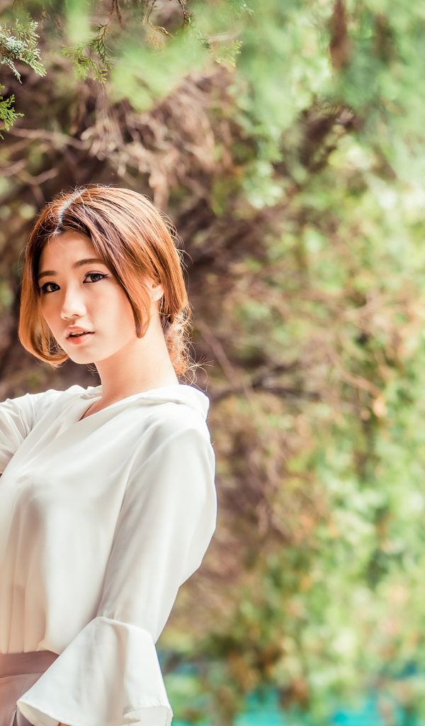 Молодая азиатка в белой блузке стоит у дерева 