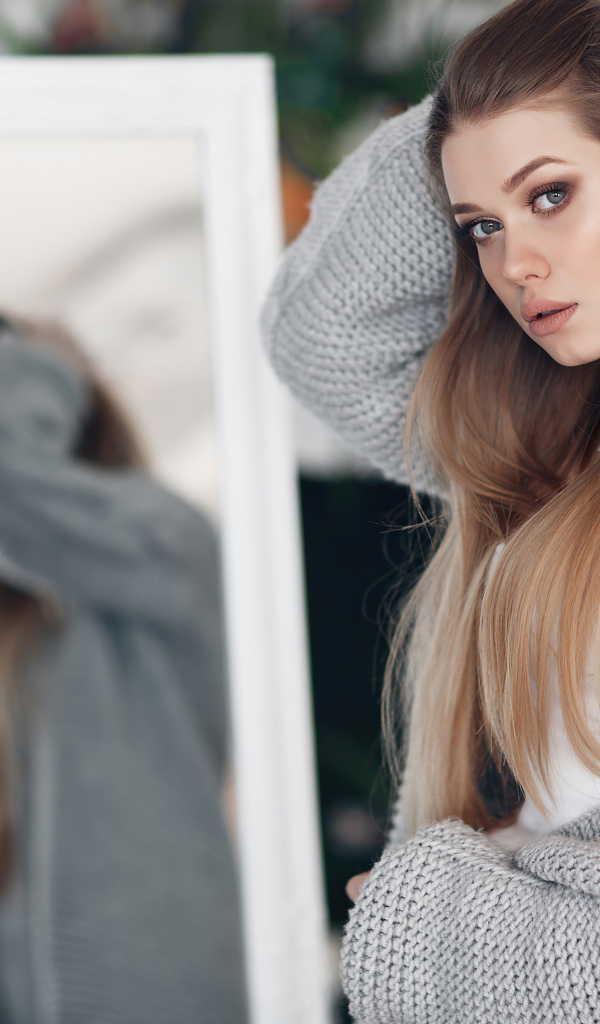 Молодая девушка в серой кофте стоит у зеркала 