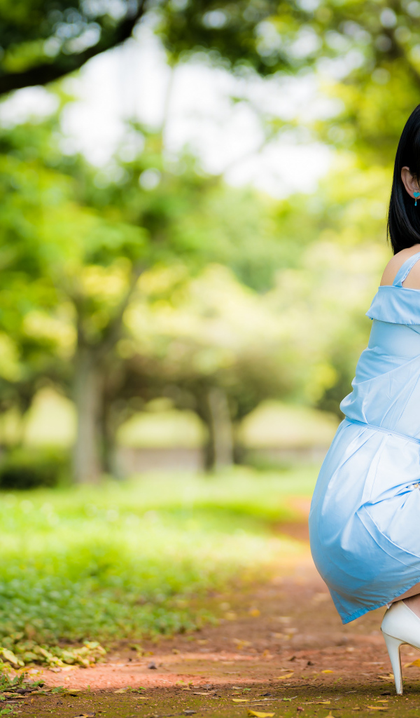 Девушка азиатка в голубом платье сидит на корточках в парке