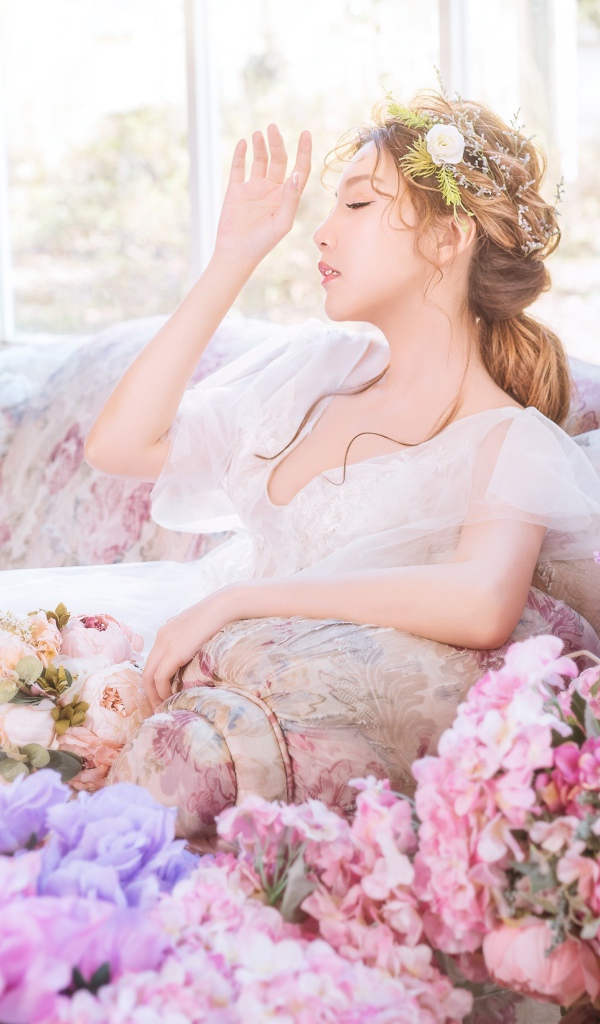 Девушка азиатка в белом платье сидит на диване в комнате с цветами