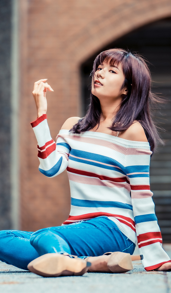 Девушка азиатка в джинсах и полосатом свитере сидит на асфальте