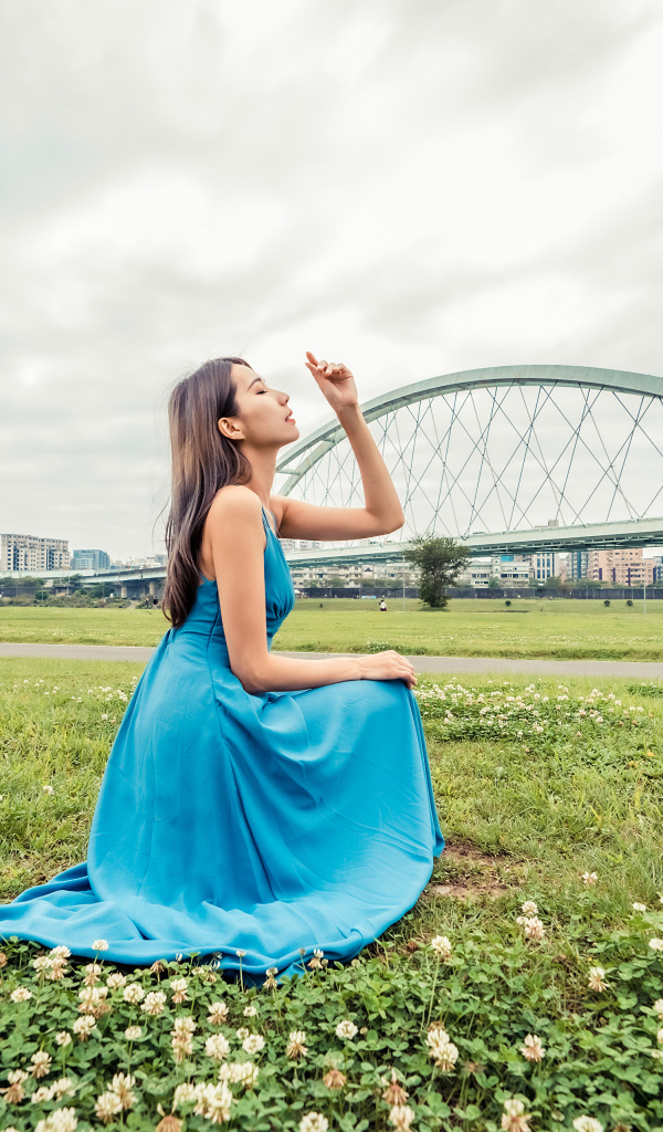 Красивая девушка азиатка в голубом платье  сидит на траве на фоне моста